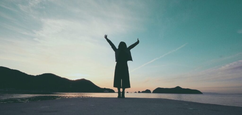 夕暮れ、あるいは夜明けの海を背景に、海岸に立つ人物のシルエット。喜びや開放感を表すように、両手を高く空に掲げている。背景には島や山の影が見え、空は穏やかなグラデーションを描いている。